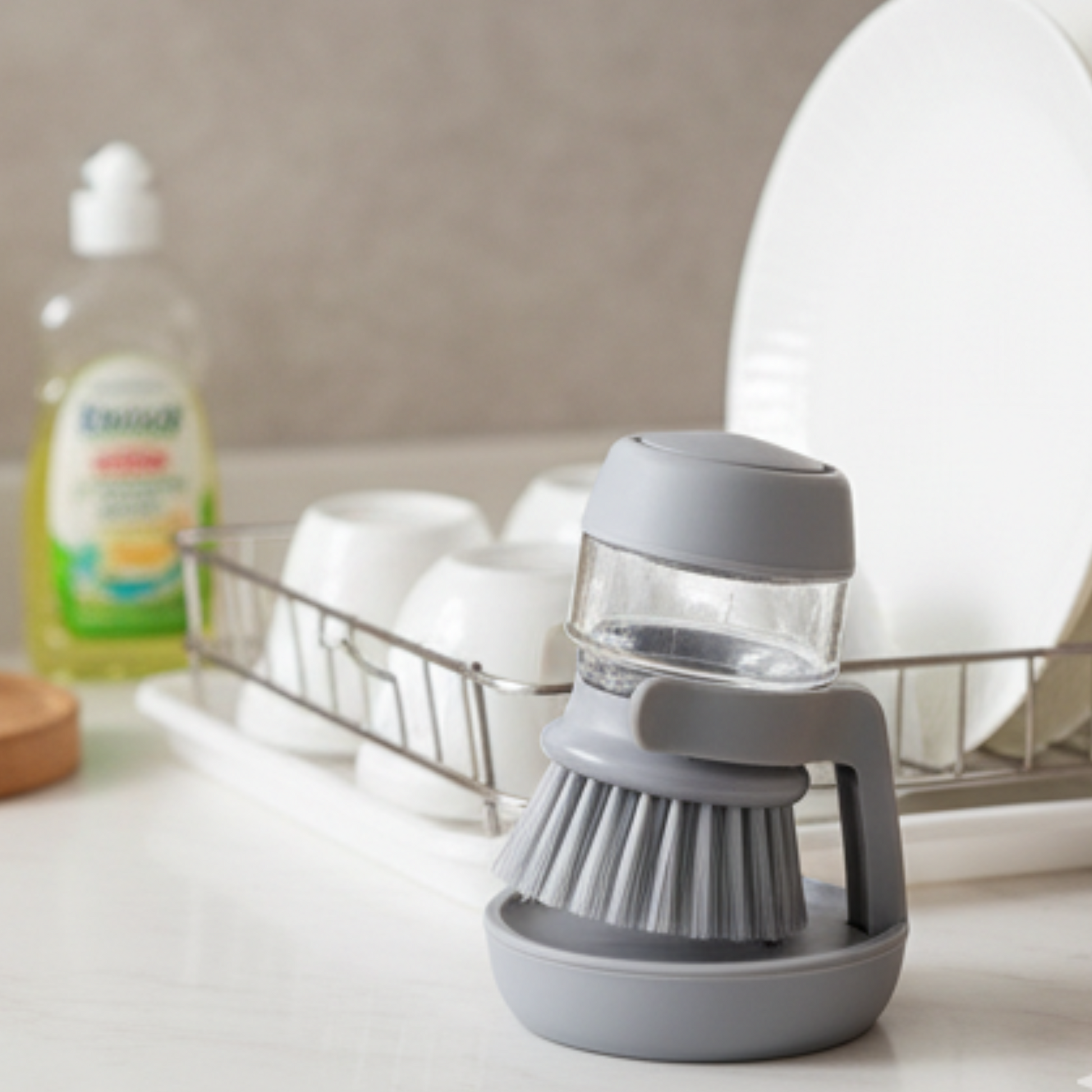 Gray dish scrubber with brush on a kitchen counter, with a dish rack and cleaning products in the background.